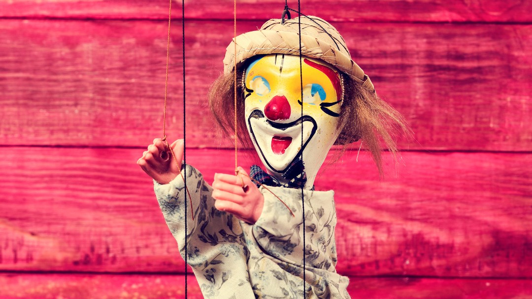 A painted clown marionette on strings against a red wooden wall.